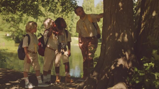 Full length shot of mid adult Caucasian male teacher and diverse group of elementary school children talking while standing under tree in forest on sunny day scouting together