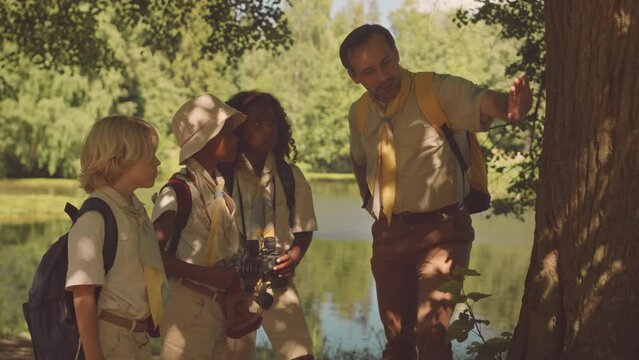 Medium shot of mid aged Caucasian male scout leader showing small group of kids around forest flora, standing by huge tree trunk on sunny day
