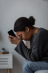 Fototapeta premium Side view of young multiracial woman with mental problem holding cellphone while sitting on bed at home.