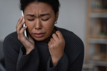 Crying multiracial woman talking on smartphone with helpline at home.