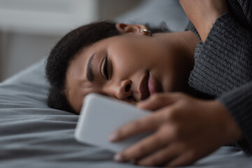 Frustrated multiracial woman using blurred cellphone while lying on bed at home.