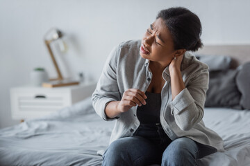 Tired multiracial woman touching neck and suffering from pain in bedroom at home.