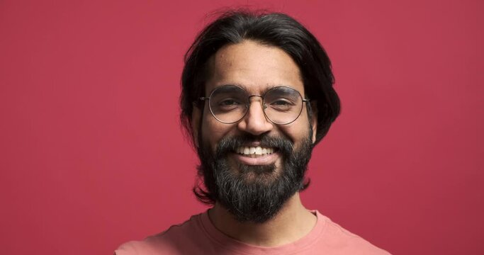 Closeup portrait of charming young man with beard and eyeglasses smiling and showing v sign of peace isolated against red background