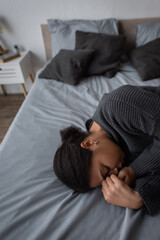 Depressed multiracial woman in knitted sweater lying on bed in blurred bedroom.