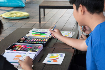 A boy in casual dark blue clothes is practicing drawing and painting by using small watercolor rectangle bars in a paint box on a table on terrace of his summer vacation.