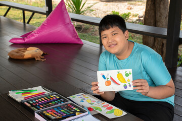 A boy in casual blue clothes is showing his drawing and painting which he used small watercolor...
