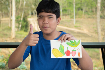 A boy in casual dark blue clothes is thumbing up and showing his drawing and painting which he used...