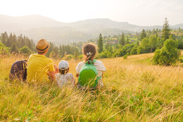Naklejka na ściany i meble Happy young family sitting back view mountain family nature vacation green travel together nature child mountain kid parents child landscape kid. Green weekend family looking at mountain travel nature