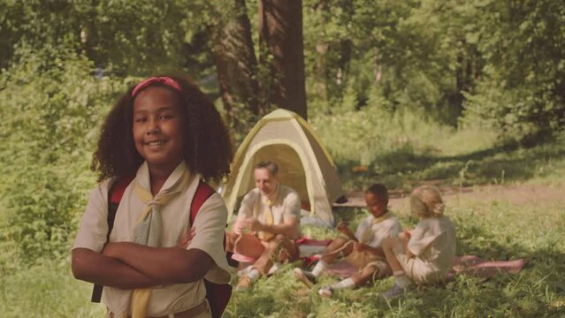 Medium slowmo portrait of smiling African American girl scout in uniform posing for camera with hands folded standing on sunny forest glade, camping with troop and leader in summer