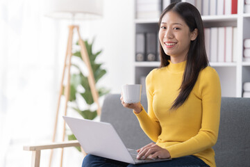Young Asian woman sitting on sofa holding Teacup and notebook and check email, in the leaving room.
