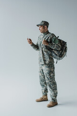 military man in cap and army uniform standing with backpack and holding smartphone on grey.
