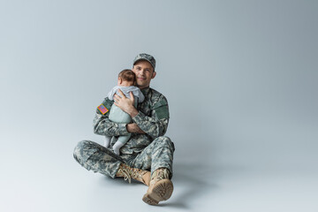 USA soldier in uniform holding in arms newborn boy while sitting on grey.
