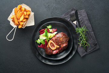 Steaks Osso Buco. Sliced beef grilled Osso Buco or porterhouse meat steak with garnished with salad and french fries on black marble board on old wooden background. Top view. Mock up.