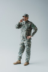 full length of serviceman in cap and army uniform talking on smartphone and standing on grey.