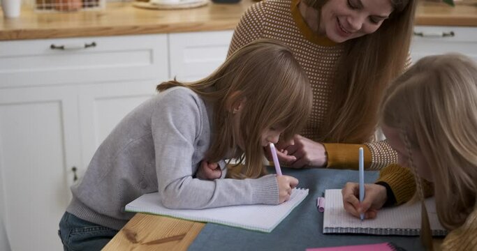 Slow motion shot of smiling young mother assisting daughters in writing homework in notebooks on table at home