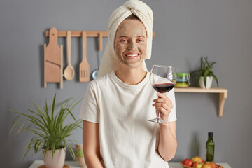 Joyful satisfied relaxed young woman posing in kitchen with towel on head drinking wine in the morning enjoying weekend smiling to camera.