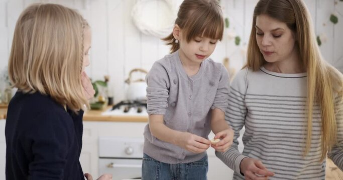 Caucasian blond young mother teaching cute daughters to make cookies with fresh dough while standing in kitchen during weekend