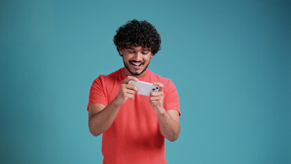 Excited happy young indian man gamer player using smartphone, playing game in mobile videogame app or watching digital video stream holding cell phone isolated on blue studio background. - obrazy, fototapety, plakaty