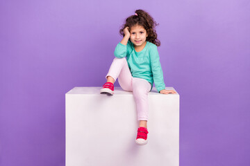 Photo of adorable sweet little child dressed teal shirt sitting white cube podium empty space isolated violet color background