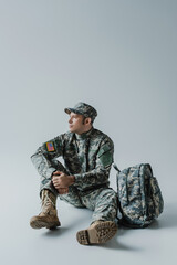 American soldier in army uniform with national flag sitting near backpack on grey.