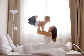 Beautiful blond child and his mother, playing at home in the morning in bed, smiling, laughing and...