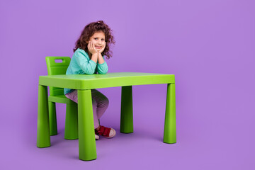 Full length photo of shiny cute little child dressed teal shirt sitting table studying empty space isolated purple color background