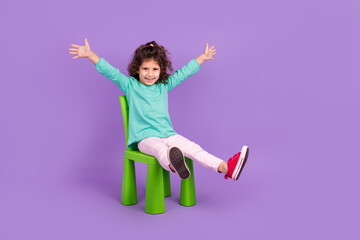 Full length photo of excited funky small kid wear turquoise shirt sitting chair rising hands arms...