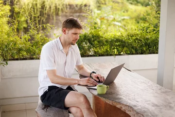Stoff pro Meter Young digital nomad working remotely on his laptop in Bali. Freelancer Man in workation video call. Travel and remote online work. Outdoor in the garden desk for  remote workers someone abroad.  © Girts