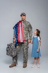 army soldier in uniform with flag of United States of America holding backpack and standing with daughter on grey.