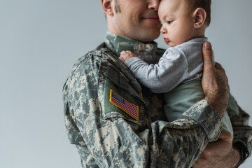 American serviceman in military uniform holding in arms infant son isolated on grey.
