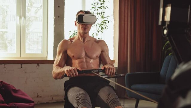 Strong young man with relief, muscular, fit, shirtless body training at home stationary rowing machine in VR glasses. Modern training. Sportive lifestyle, body and health care, fitness, health concept