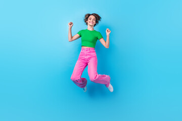 Full length photo of cute lucky woman dressed green top jumping high rising fists isolated blue color background