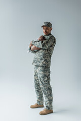 full length of army serviceman in uniform and cap holding newborn son on grey.