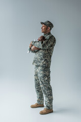 full length of pleased army serviceman in uniform and cap holding newborn son on grey.