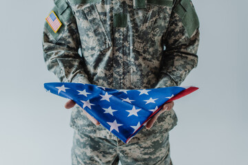 cropped view of American soldier in uniform holding folded flag of United States during memorial day isolated on grey.