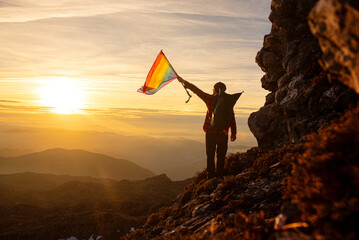 Hiker man with backpack waving a rainbow lgbt pride flag on the mountain at sunset. Concept of...