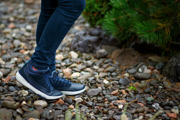 Blue leather sneakers with a white sole and blue laces, shod on the girl's feet, stand on pebbles in the garden against the backdrop of coniferous bushes