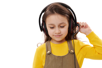 Mischievous little kid girl, primary school student, first grader with headphones, smiling over white isolated background. Smart pupil enjoys online lesson on audio headset. Distance education concept