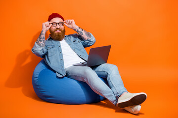 Full length photo of smart clever man wear jeans shirt sitting bean bag working device empty space isolated orange color background