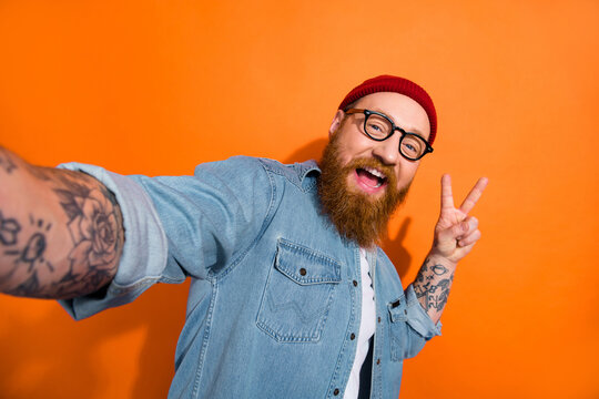 Photo of funky positive guy dressed denim jacket eyeglasses showing v-sign taking selfie isolated orange color background