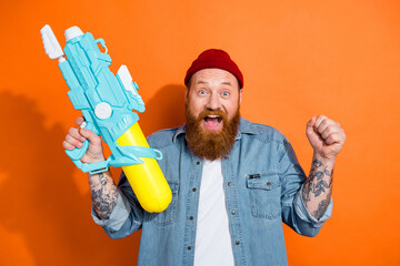 Photo of excited lucky guy dressed denim jacket rising water gun fist winning game isolated orange color background