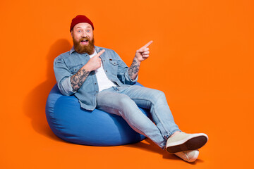 Full length photo of positive excited man wear jeans shirt sitting bean bag pointing empty space isolated orange color background