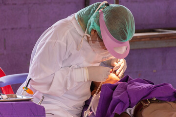 A dentist wearing a white shirt is doing dental work for a male student lying on a bed at the school grounds.