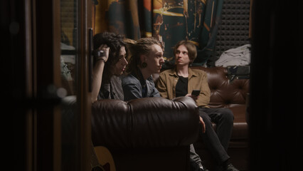 Musical rock band sits on sofa. Young male musicians, artists, performers listen to finished recorded songs for new album in professional sound and music recording studio. Concept of music production.
