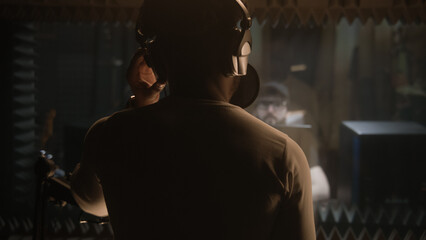 African American vocalist sings composition into microphone in soundproof room. Experienced audio engineer or producer works with singer in sound recording studio. Music production concept. Back view.