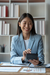 Confident smiling asian businesswoman sitting at desk while pressing calculator to calculate earnings Income from graph document data Chart showing financial business growth at the office.