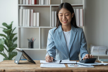 Confident smiling asian businesswoman sitting at desk while pressing calculator to calculate earnings Income from graph document data Chart showing financial business growth at the office.