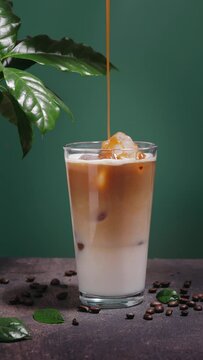 Pouring caramel sauce into a glass with iced coffee on dark teal background with coffee leaves
