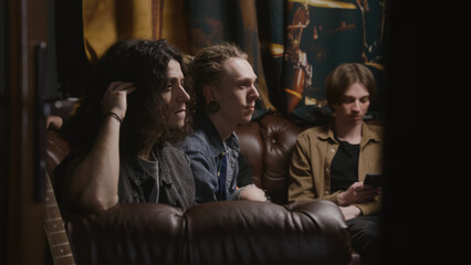 Musical rock band sits on sofa. Young male musicians, artists, performers listen to finished recorded songs for new album in professional sound and music recording studio. Concept of music production.