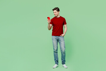 Full body young smiling brunet caucasian man he wears red t-shirt casual clothes hold in hand use mobile cell phone isolated on plain pastel light green background studio portrait. Lifestyle concept.